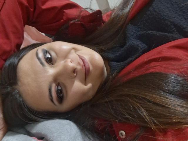 KatherinLatin, lying on her back with long brown hair, red shirt, and black top, smiling with a nose ring, brown eyes, and subtle makeup.