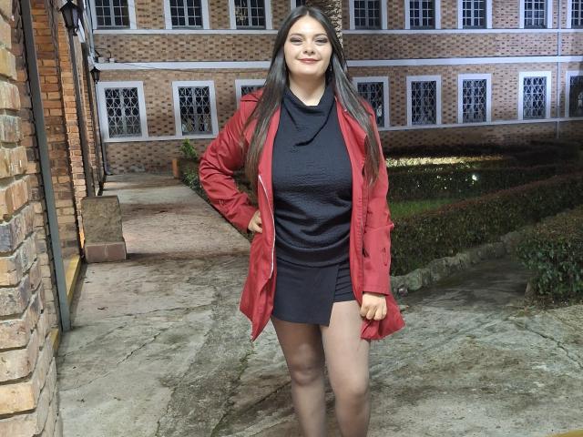 Katherin Latin tiene el pelo largo y castaño, una chaqueta roja, una camisa negra y una falda corta negra. Está de pie con una mano en la cadera, mostrando su trasero..