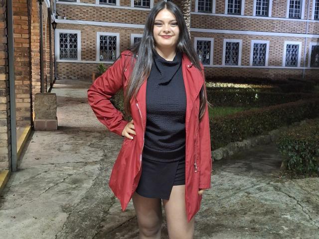 KatherinLatin with long dark hair, smiling, wearing a red coat over a black turtleneck and black mini-skirt, standing on a stone path, hands on hips, showing her ass.