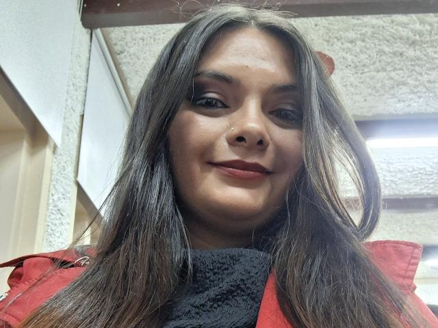 Katherin ha lunghi capelli castani, un piccolo piercing al naso, indossa una giacca rossa, un maglione nero a collo alto e ha un leggero sorriso. Le sue natiche non sono visibili. Sta guardando verso il basso la fotocamera..