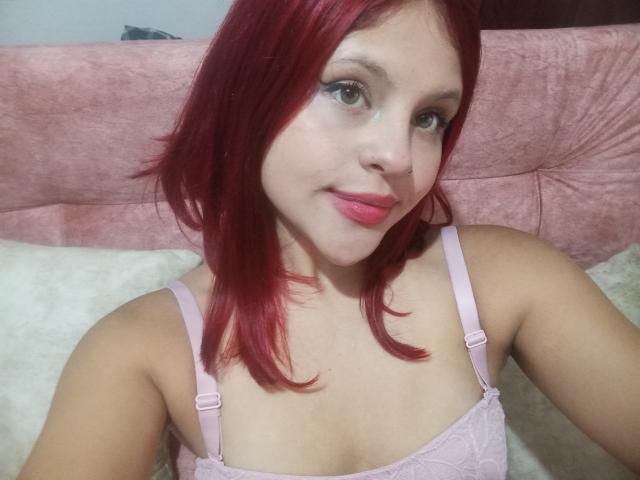SophieHill with red hair, light skin, and pink lipstick, wearing a pink lace bra, smiling slightly, showing her small tits, and green eyes.