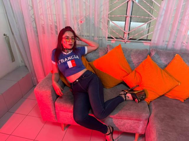 QueenScarletSecret com cabelo ruivo, óculos, cropped azul com a inscrição "FRANÇA", jeans pretos, almofadas laranjas, sofá cinza e saltos altos pretos, inclinada para trás, com a mão no cabelo, iluminação vermelha..