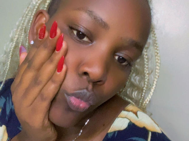 SweettCaandy, avec une peau brun foncé, des ongles rouges et des tresses blondes, les lèvres boudeuses, une main sur la joue, portant un haut fleuri, un boucle d'oreille en diamant et une expression sérieuse..