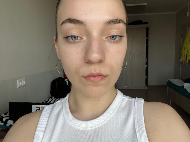 ChasidyCockrin has pale skin, green eyes, and a serious expression. She's wearing a white tank top, hoop earrings, and has her hair pulled back. Her nose is slightly upturned.