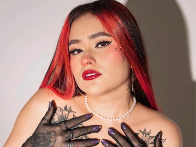 Marystixr with red and black hair, wearing a pearl necklace, black lace gloves, purple nails, and tattoos on her chest, looking at the camera with red lipstick.