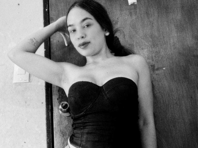 Fotografia in bianco e nero di CloeSantos con capelli scuri, che indossa un abito nero senza spalline, con una mano tra i capelli, piccoli orecchini, un tatuaggio sul braccio sinistro, in piedi di fronte a una porta di legno..