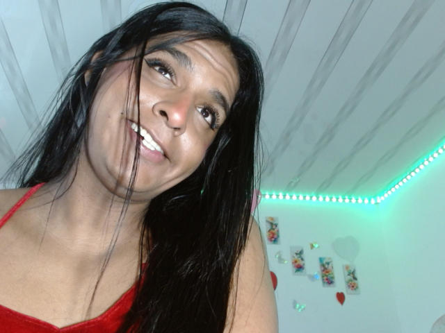 IdiiaLatina mit langen, schwarzen Haaren, die ein rotes Tanktop trägt, lächelt in die Kamera, wobei ihr Kopf leicht geneigt ist, und zeigt ihre Zähne. Sie befindet sich in einem hellen Raum mit LED-Streifen..