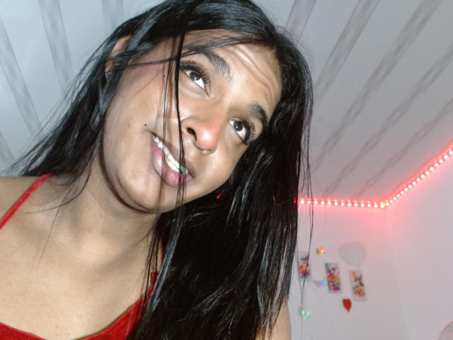 IdiiaLatina with long black hair, dark brown eyes, and a nose ring, smiling slightly, wearing a red top, her face in the foreground, and red LED lights in the background.