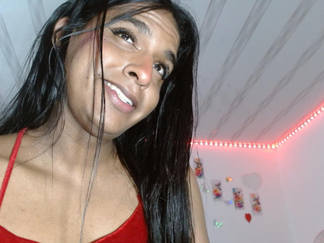 IdiiaLatina with long black hair, brown eyes, and a smile, wearing a red tank top, showing her tits, in a brightly lit room with red LED strip lights.