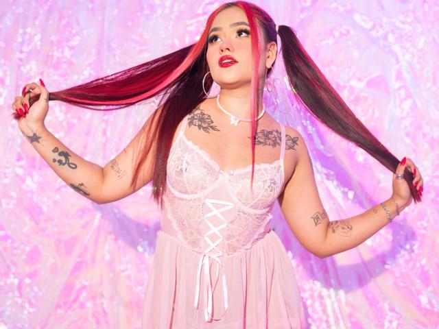 Marystixr with red and black pigtails, wearing a pink lace babydoll, has tattoos on arms and chest, red lipstick, and holds pigtails, wearing a white choker.