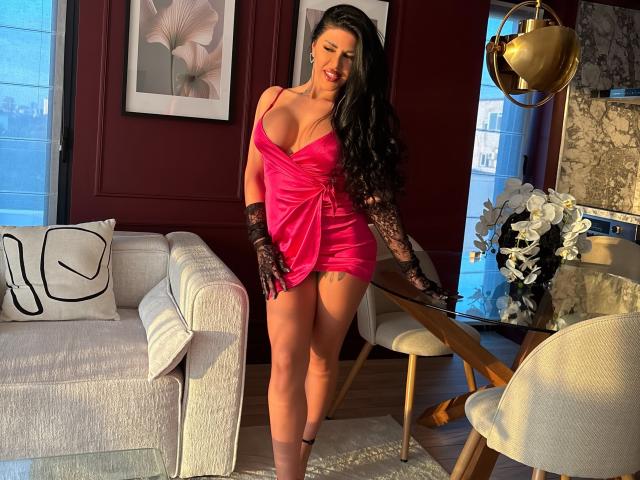 GallienneGray mit großen Brüsten in einem pinkfarbenen, schulterfreien Minikleid, schwarzen Spitzenhandschuhen und langen schwarzen Haaren, stehend in einem Raum mit einem weißen Sofa und einem Glastisch..