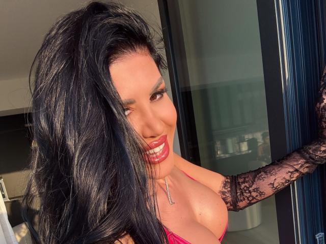 GallienneGray con lunghi capelli neri, sorridente, rossetto rosso, braccio con pizzo, reggiseno rosa, che mostra un seno prosperoso, luce solare, porta, collana, che guarda la fotocamera..
