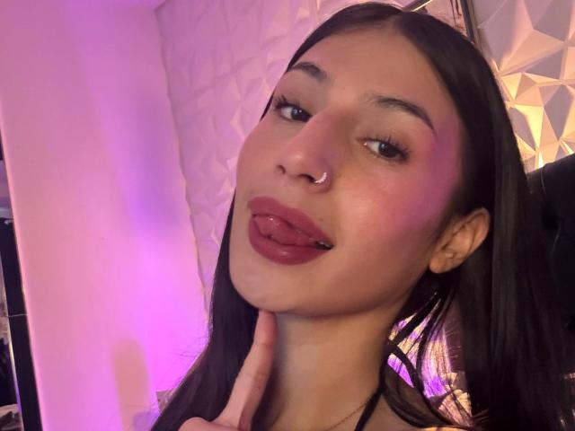 SophieBeaumont with long black hair, nose ring, and pinkish-purple skin, sticking out her tongue, looking at the camera, with her finger near her chin.