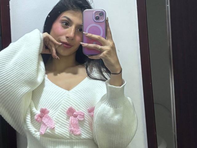 Sophie Beaumont met een roze blos, lang donker haar, een witte gebreide trui met roze strikjes, die een roze telefoon vasthoudt, een spiegelzelfportret, hand op de wang, zachte glimlach, witte muur, donkere deur..
