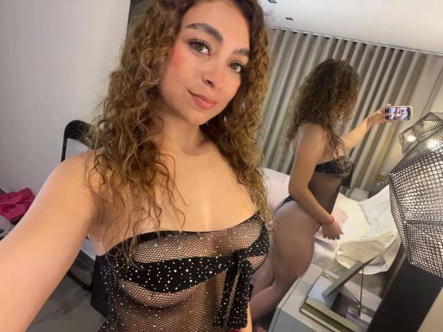 Arianna Blake, met krullend bruin haar, draagt een zwarte kanten top die haar borsten onthult, en maakt een selfie in een spiegel. Haar achterkant is zichtbaar in de reflectie..