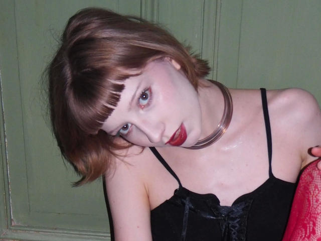Alexa, séduisante avec une peau pâle, des cheveux bruns courts, des yeux bleus, du rouge à lèvres rouge, une robe noire, un collier brillant, penchée en avant, montrant ses seins et sa main rouge, regardant l'appareil photo..