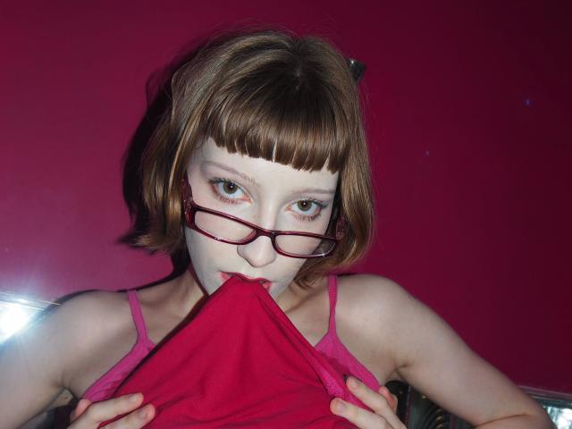 Alexa, sexy e attraente, con capelli castani corti, occhiali rossi e reggiseno rosa, morde la sua biancheria intima rossa, mostrando il suo viso pallido, con lentiggini e occhi verdi..