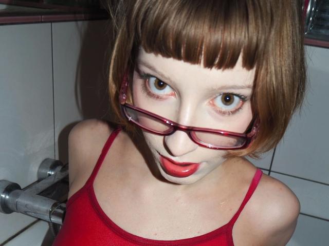 Alexa, très sexy, avec des cheveux courts bruns, du rouge à lèvres rouge et des lunettes rouges, regardant vers le haut avec de grands yeux bruns, portant un débardeur rouge, dans une salle de bain carrelée, montrant ses seins..