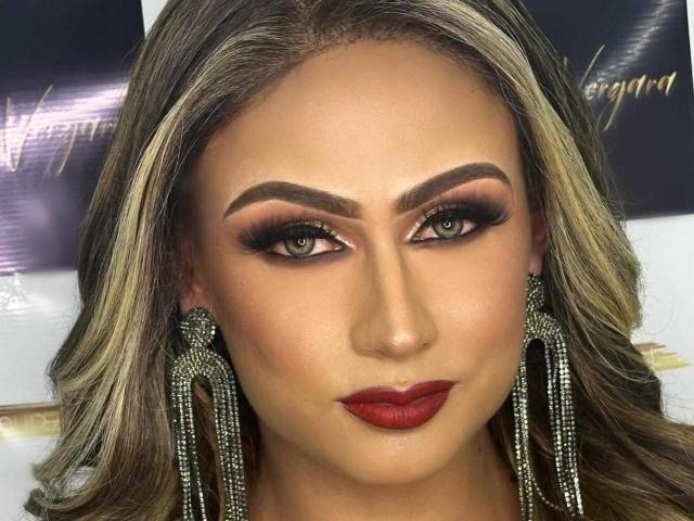 QueenMelodyX has medium skin, green eyes, dark red lipstick, and dramatic makeup. She's wearing large, sparkly earrings and has blonde, wavy hair with dark roots.