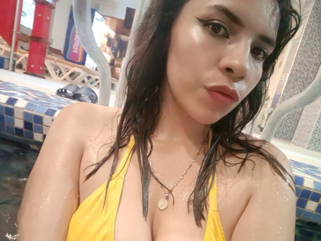 VioletaStone, con el cabello castaño mojado, un top de bikini amarillo, mostrando sus pechos, en una piscina, llevando un collar, mirando a la cámara con la piel brillante y un maquillaje sutil..