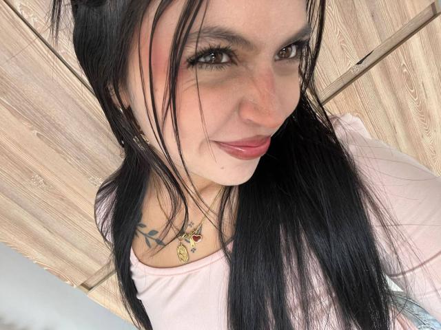 NyraVanel tem cabelos pretos longos, olhos castanhos e um sorriso discreto. Ela está vestindo uma blusa rosa com um ombro de fora, revelando uma tatuagem em seu ombro direito. Seus seios estão parcialmente visíveis..