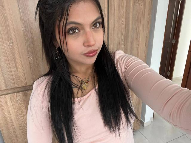 NyraVanel mit langen, schwarzen Haaren, braunen Augen, einem pinkfarbenen Oberteil mit langen Ärmeln, einer goldenen Halskette und rotem Lippenstift, die ein Selfie macht. Brüste nicht sichtbar, Vulva nicht sichtbar, Hintern nicht sichtbar..