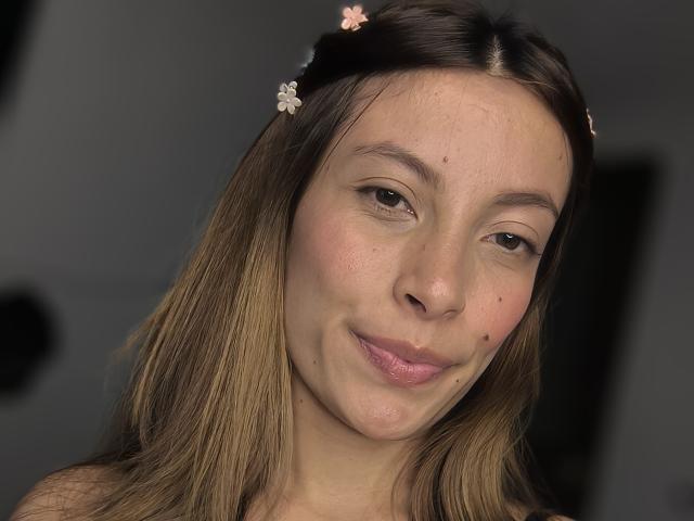 KiaraWiild a la peau brun clair, les yeux marron, et une petite tache de beauté sur sa joue droite. Elle sourit, avec du rouge à lèvres rose, et porte de petites pinces à cheveux en forme de fleurs roses et blanches..