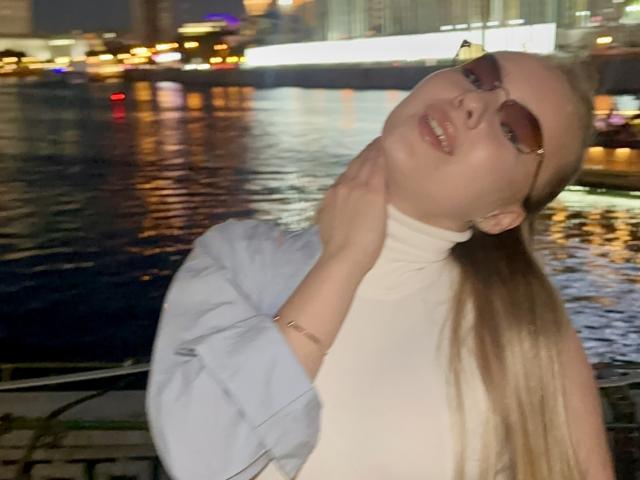 Karleen Carthen mit langen blonden Haaren, die eine Sonnenbrille trägt, ein weißes Rollkragenoberteil und ein hellblaues Hemd, ihr Kopf ist leicht nach hinten geneigt, sie lächelt, ihre Hand liegt auf ihrem Hals, vor einem dunklen, spiegelnden Gewässer in der Nacht..