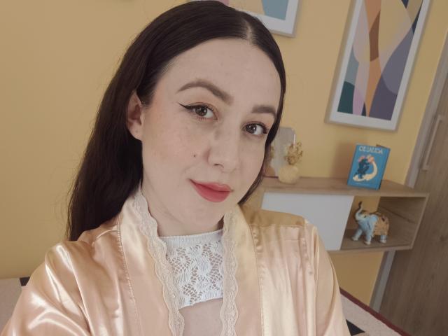Maby Legrand, avec sa peau pâle, ses cheveux foncés et son rouge à lèvres rose, porte une robe en satin rose brillante avec de la dentelle blanche. Elle a un maquillage discret et un joli nez, et elle sourit doucement..