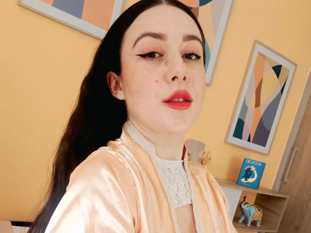 MabyLegrand, met bleke huid, zwart haar, rode lippenstift en een roze satijnshirt met kant, kijkt naar de camera met halfgesloten ogen. Ze heeft een klein beauty mark..