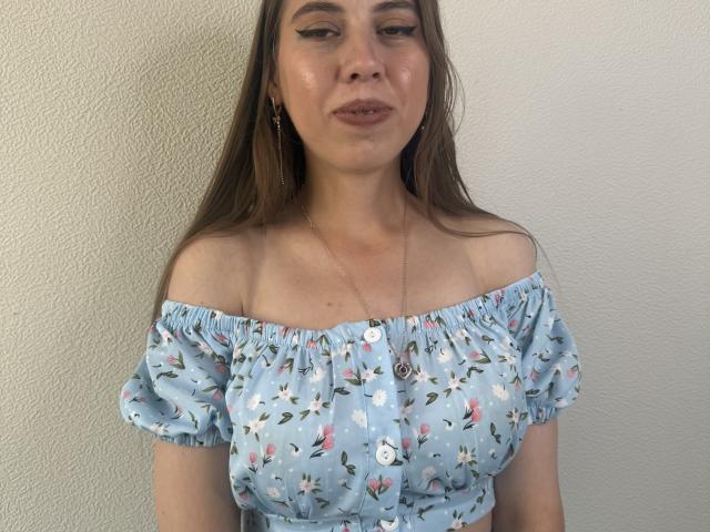 Shelli Wainman, con i capelli castano chiaro, indossa un abito floreale blu con una spalla scoperta, mostrando piccoli seni, una leggera abbronzatura e un sorriso sicuro, sullo sfondo di un muro bianco..
