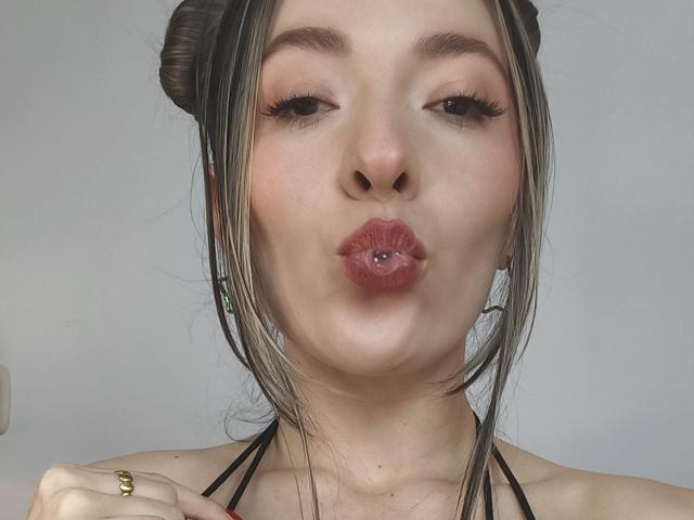 TaniaSandersX, con le labbra tumefatte, un anello d'argento al labbro e i capelli biondi raccolti in due chignon, fa un'espressione civettuola. Indossa un top nero con spalline e un anello dorato..