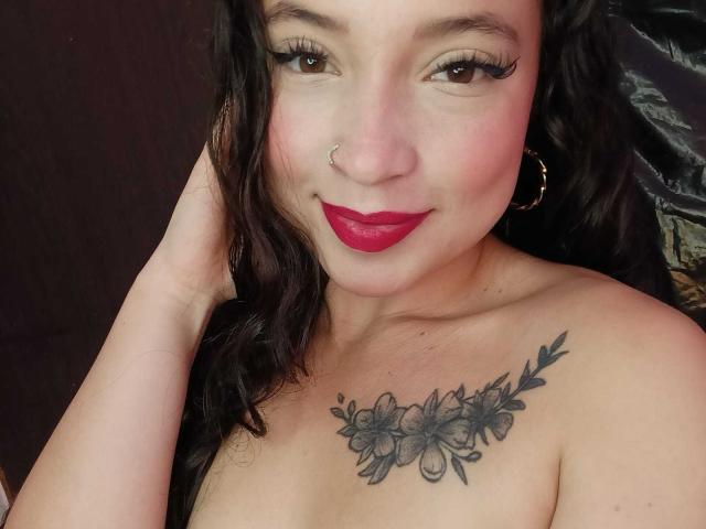 MiaAlessia has dark wavy hair, red lipstick, a nose ring, hoop earrings, and a floral tattoo across her tits. She's smiling with long eyelashes and a hand in her hair.
