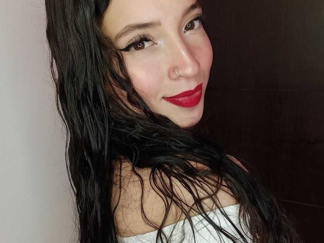 MiaAlessia has wet black hair, red lipstick, and a nose ring, with her tits barely covered by a white strap. She's smiling at the camera.