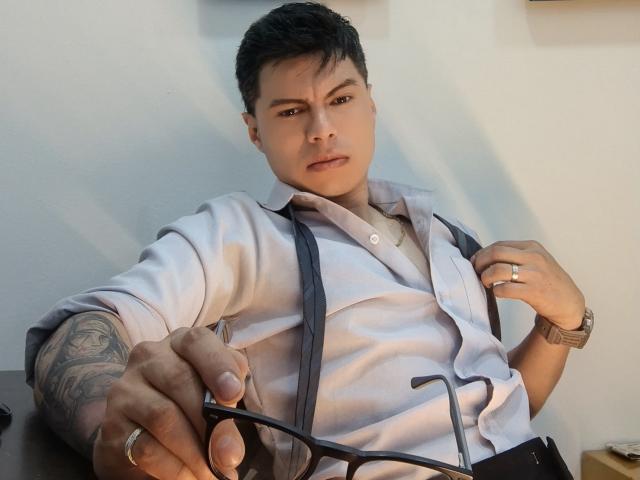 Aquiles Taylor, musculoso, con tatuajes, cabello negro, camisa blanca, tirantes grises, sosteniendo gafas, anillo en el dedo, reloj en la muñeca, expresión seria, recostado, interior, iluminación suave..