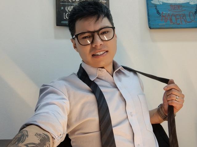AquilesTaylor with short black hair, glasses, and tattoo on forearm, holding black tie over white shirt, smiling at camera, right hand on tie, left arm visible.