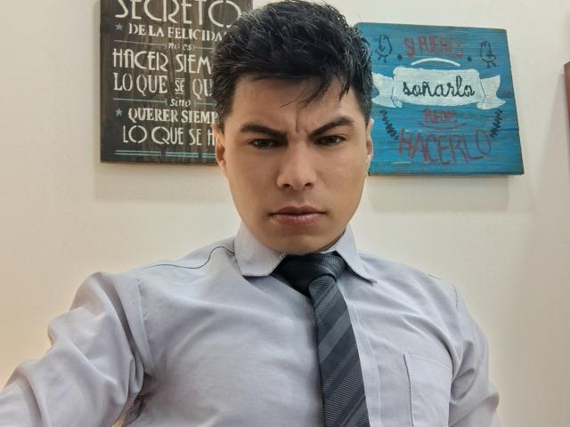 AquilesTaylor a une expression sérieuse, il porte une chemise bleu clair et une cravate rayée foncée, avec des cheveux noirs courts, sur une photo de lui-même prise avec un angle légèrement vers le bas..