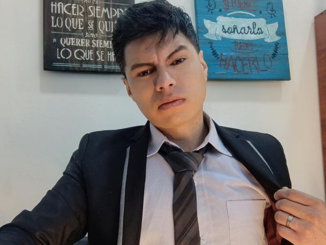 AquilesTaylor, a serious-looking dude with short black hair, wearing a black suit, white shirt, and gray tie, holding his jacket with a ring on his right hand.