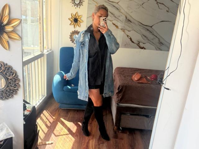 Elena Saenz, con una chaqueta vaquera, una camiseta negra y pantalones cortos, y botas negras hasta la rodilla, está tomando una foto de sí misma en un espejo, mostrando sus glúteos, en una habitación iluminada por el sol con una silla azul..