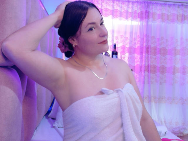 VaaleryStark with a towel wrapped around her tits, arm raised, brown hair in a bun, silver necklace, pink and purple lights in the background, looking slightly to the side.