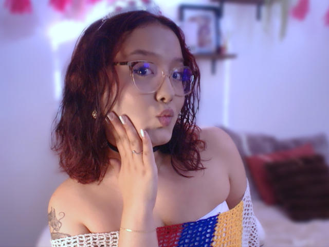 SophiaBrowm with red hair, glasses, and a colorful off-shoulder top, pouting with hand on cheek, showing a tattoo on her left arm. Her tits are visible.
