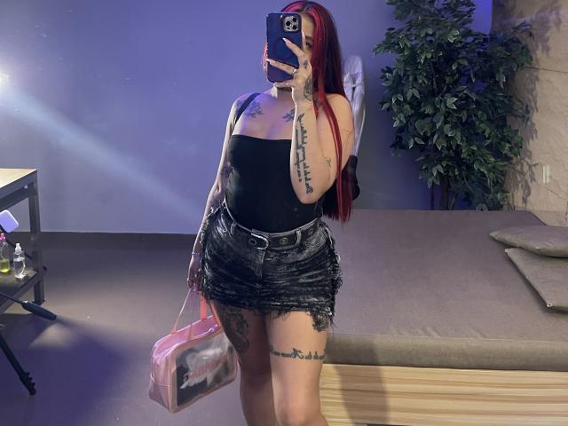 Marystixr mit roten Haaren, einem schwarzen Top ohne Träger und einem abgenutzten schwarzen Rock, hält ein Handy für ein Selfie vor dem Spiegel, hat Tattoos an Armen und Oberschenkel, eine pinke Handtasche in der Hand, violette Beleuchtung..
