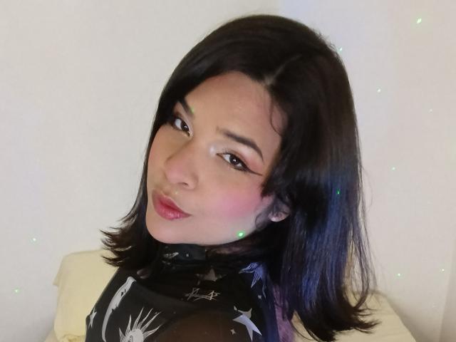 Antonela, com cabelos pretos brilhantes, blush rosa, batom vermelho e uma blusa preta com estampa de estrelas, sorrindo para a câmera, com uma expressão sexy e charmosa..