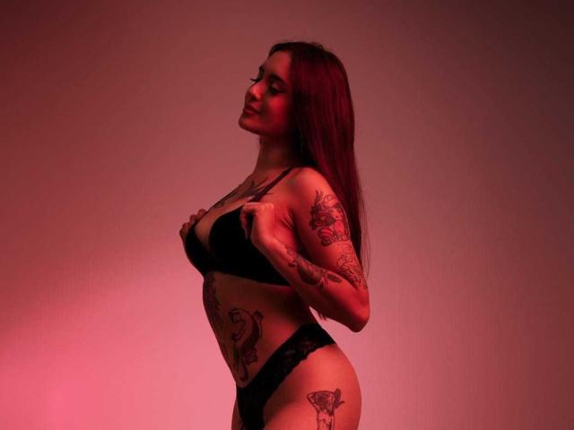 VelvetMusee con i capelli rossi, un reggiseno nero e delle mutandine nere, che si tiene i seni, con tatuaggi sulle braccia e sui fianchi, in piedi sotto una luce rossa, sorridente, che guarda di lato..