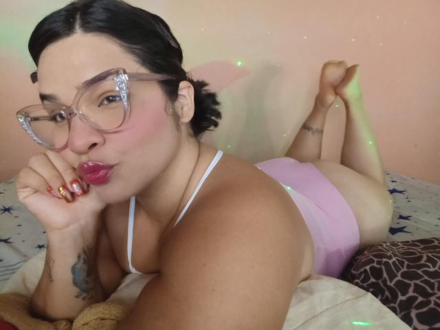 AntonelaPark, sdraiata a pancia in giù, con occhiali trasparenti, rossetto rosa e slip rosa, che mostra il sedere, con un tatuaggio sul braccio sinistro, su un letto..