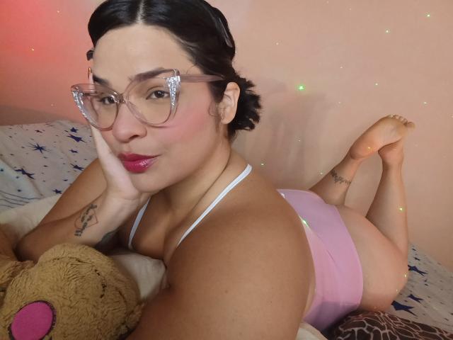Antonela, con occhiali rosa, rossetto rosa e capelli neri corti, sdraiata su un letto, indossa un reggiseno sportivo rosa, mostrando il sedere, con un orsacchiotto di peluche in primo piano..