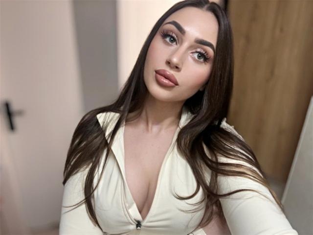 OliviAngel mit langen braunen Haaren, vollen rosafarbenen Lippen und großen grünen Augen, trägt ein weißes Top mit tiefem Ausschnitt, das ihre Brüste zeigt. Sie schaut mit einem süßen Ausdruck in die Kamera..