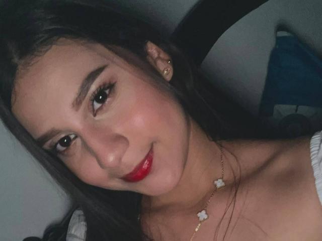 VictoriaBlue19 met rode lippenstift, donkere wenkbrauwen, lang bruin haar, draagt een gouden ketting met kleine witte bloemetjes, glimlacht zachtjes en draagt een witte top met een open schouder..