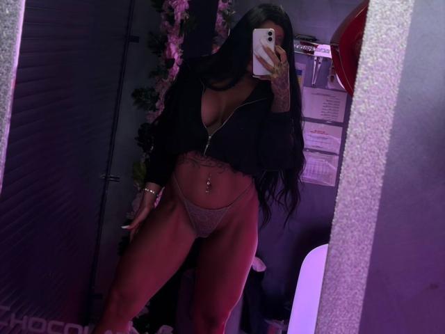 SonyaStorm, com longos cabelos pretos, vestindo uma blusa preta com zíper, calcinha de renda rosa e um piercing no umbigo, tirando uma selfie no espelho mostrando seus seios e vulva..