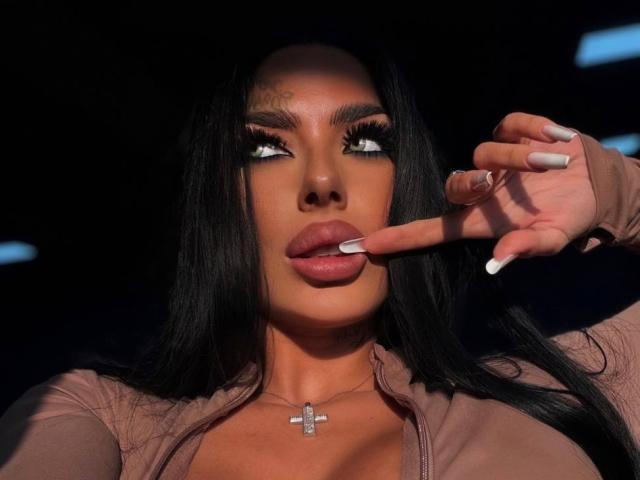 SonyaStorm with dark hair, full lips, and white nails, touching her lips with her index finger, wearing a pink top and a cross necklace, looking up.